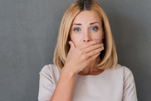 Surprised woman covering mouth with hand and staring at camera while standing against grey background