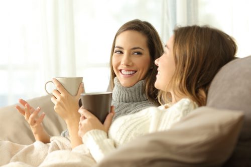 two women smiling and drinking hot drinks on a sofa