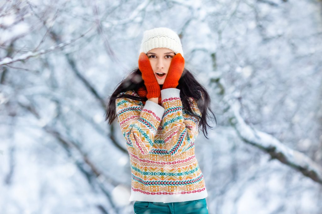 Young woman standing outside in the snow in front of a forest with red gloves on, touching her face with both hands